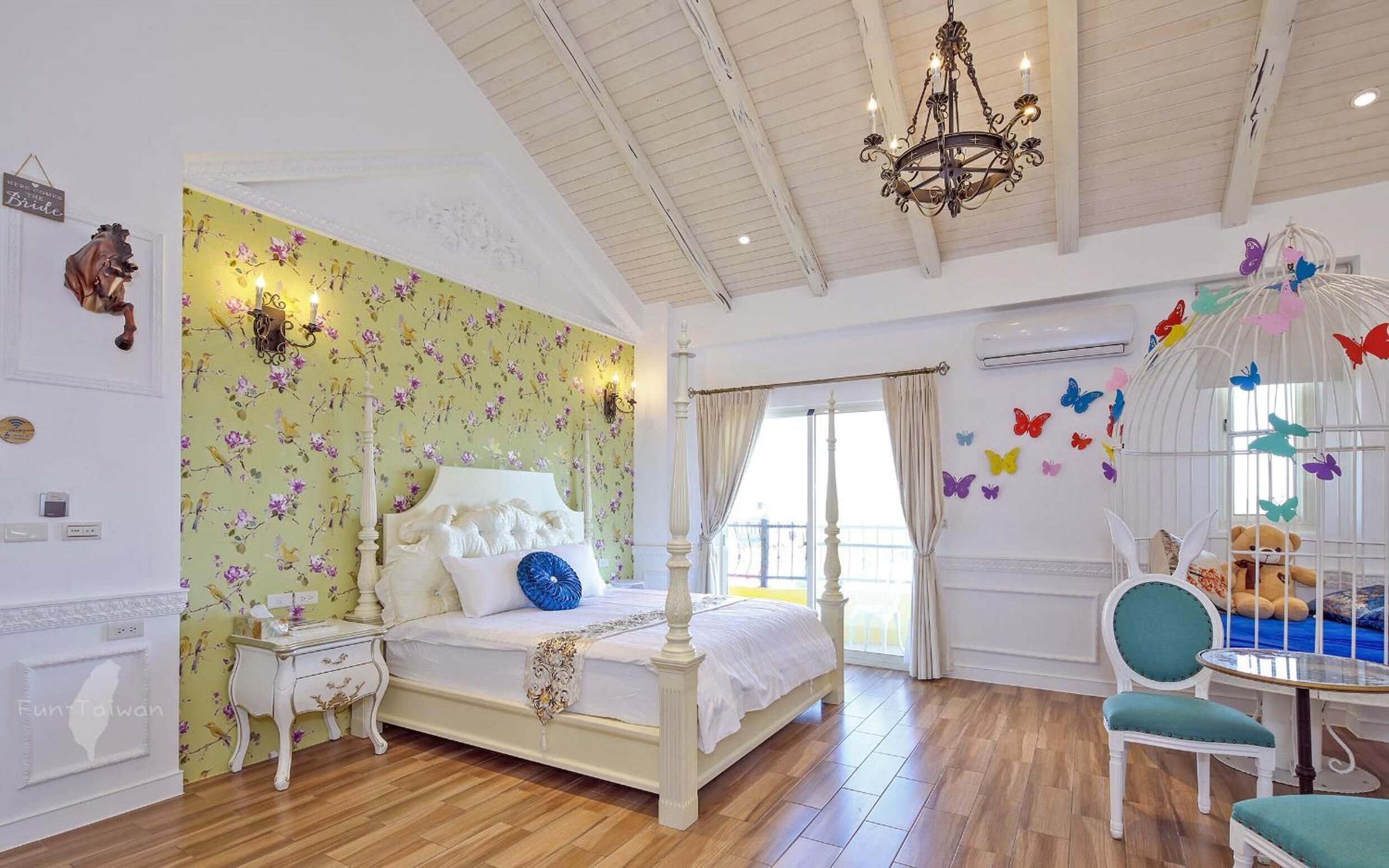 A bedroom with a floral wallpaper, a white bed, a chandelier, a window with curtains, a white chair, and a butterfly wall decoration.
