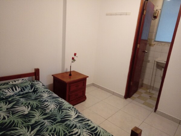 Un dormitorio con cama, una mesita de noche con un florero y una puerta que da a un baño, además de un piso de baldosas.