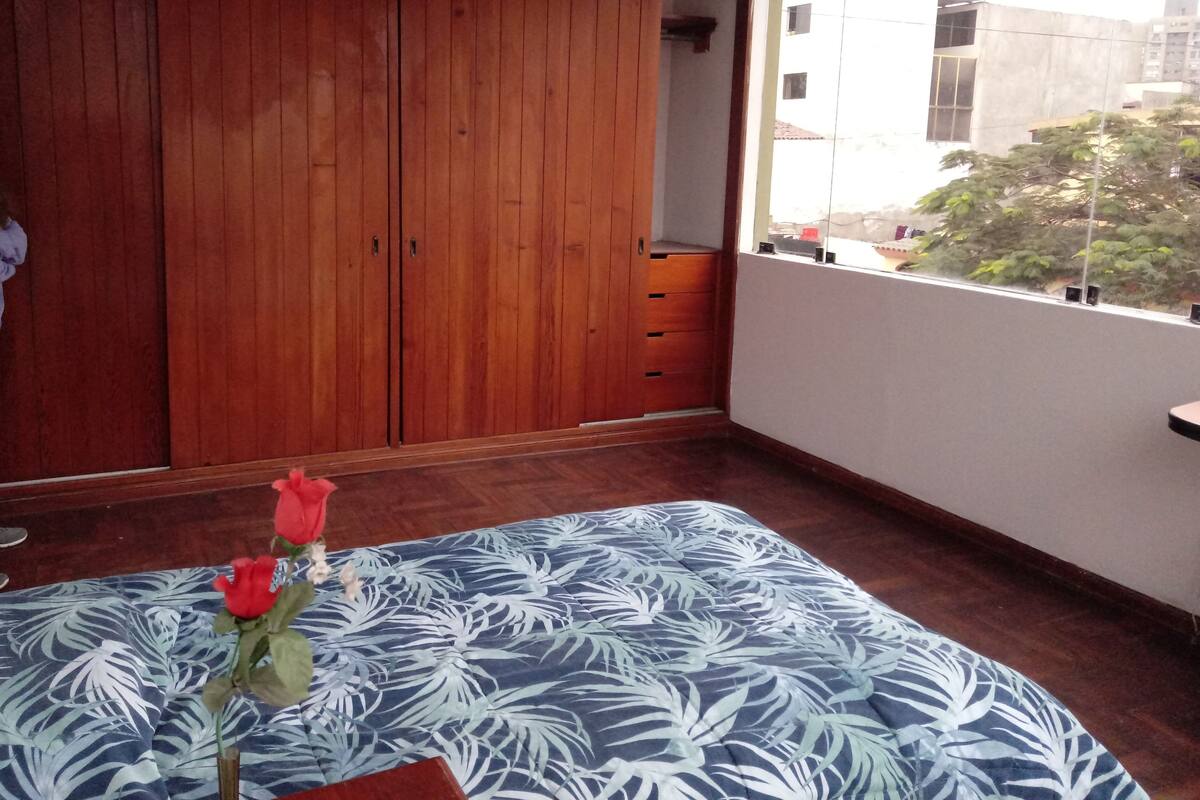 A bedroom with a large wooden wardrobe, a bed with a patterned bedspread, and a view of buildings through a window.