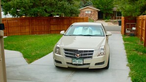 A beige Cadillac parked on a concrete driveway in a residential area with a wooden fence and a house in the background.