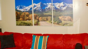 A red couch with a striped pillow and a wall-mounted triptych of a mountain landscape.