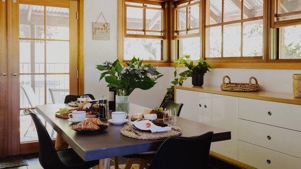 Un comedor con una mesa dispuesta para dos personas, que incluye un florero con flores, una cesta y distintos alimentos.