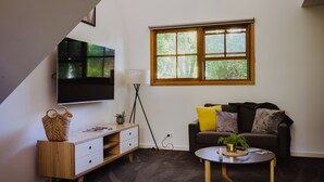 Una sala de estar moderna con una televisión de pantalla plana, un sofá con cojines, una mesa de centro y un ventanal con vistas a árboles.