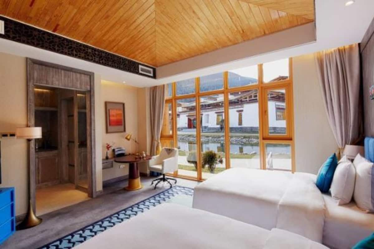 Quarto de hotel com teto de madeira, uma janela grande com vista para uma montanha, uma mesa com cadeira e duas camas.