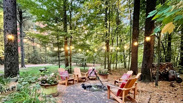 Un espace extérieur aménagé, entouré d’arbres, avec des chaises en bois et un foyer central.
