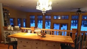 Une cuisine avec un îlot central en bois, un lustre et de grandes fenêtres donnant sur la forêt.