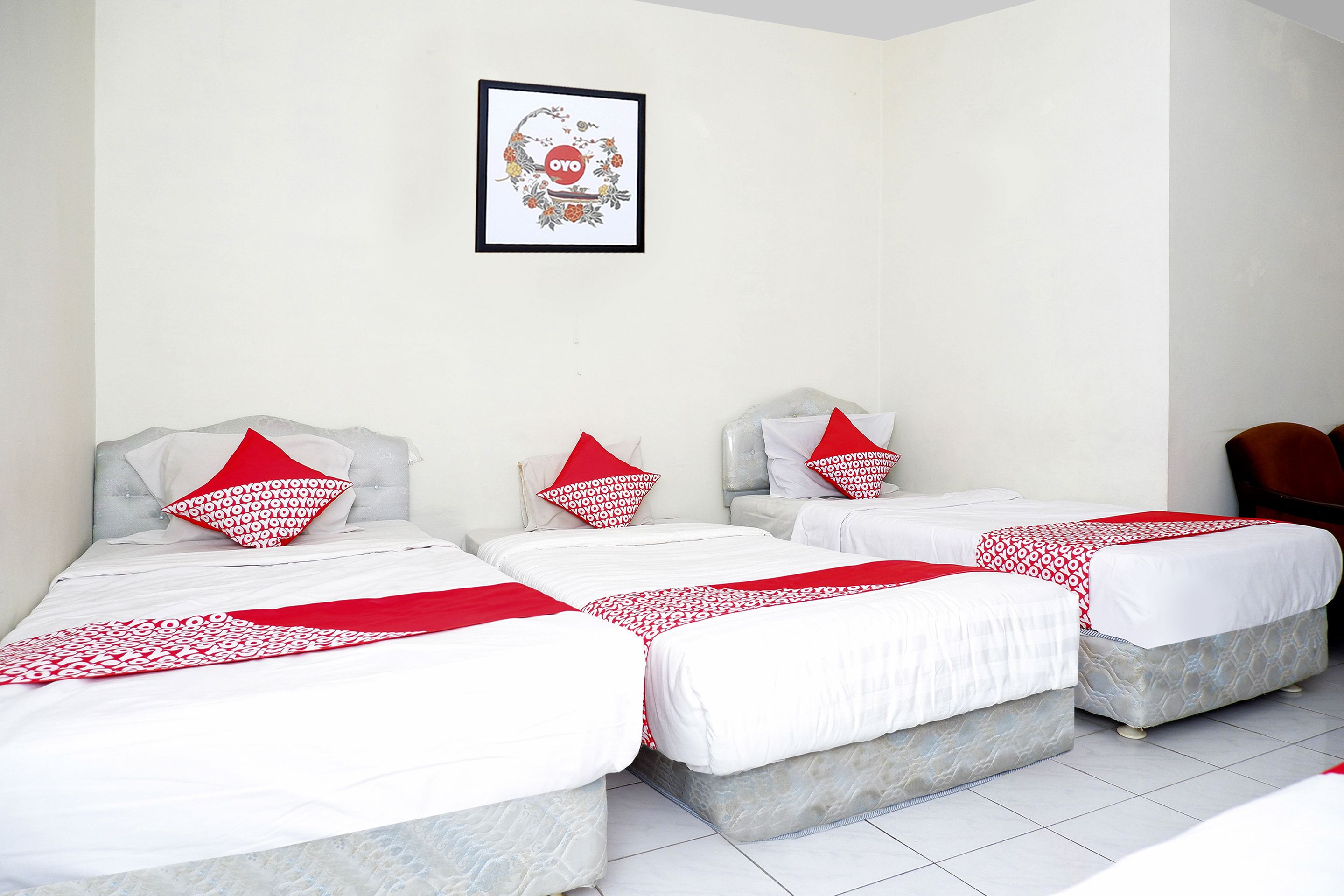 A hotel room with three beds, each with white bedding and red accents, and a framed artwork on the wall.
