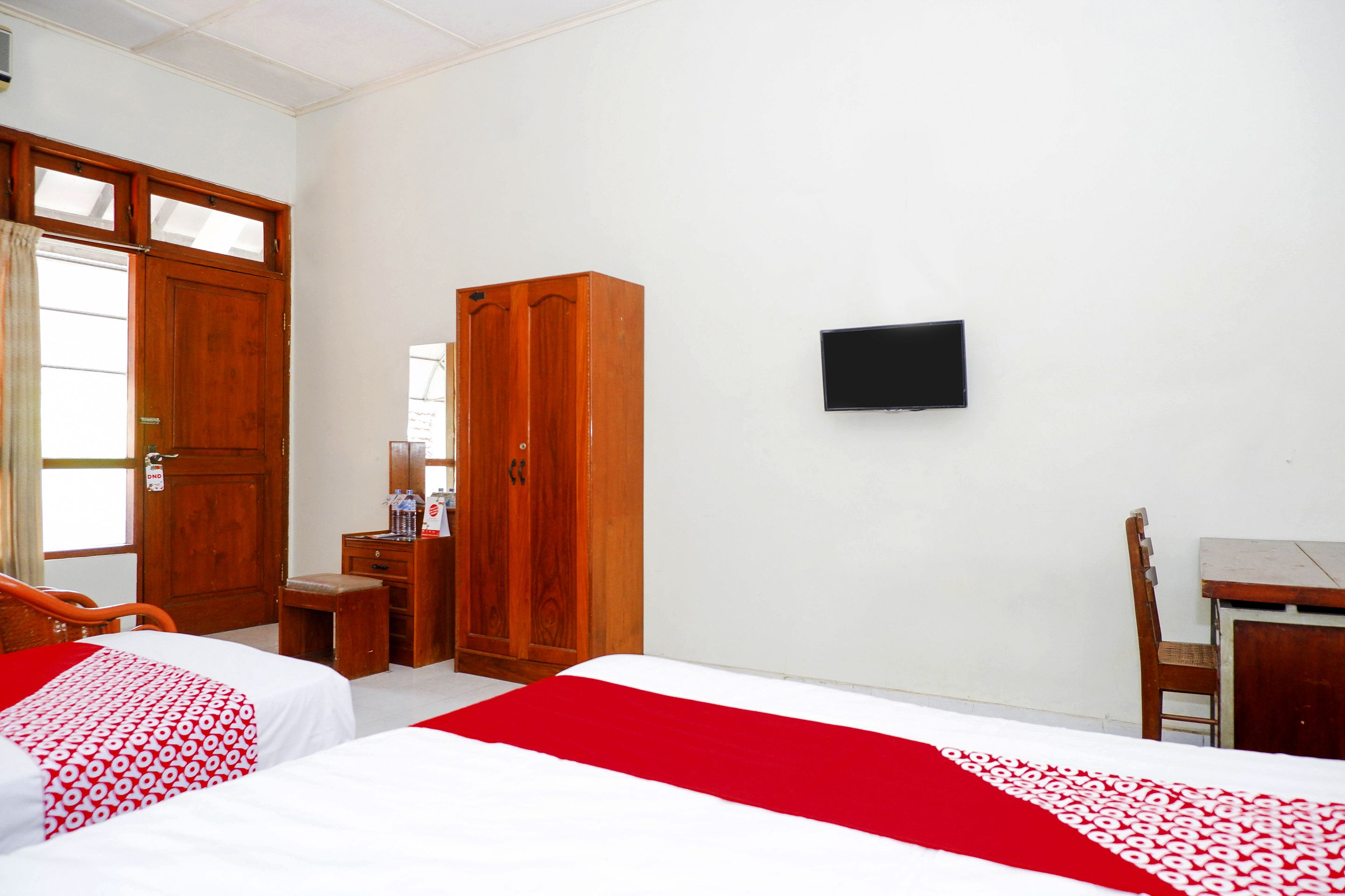 A hotel room with two beds, a wooden wardrobe, a desk with a chair, and a flat-screen TV on the wall.