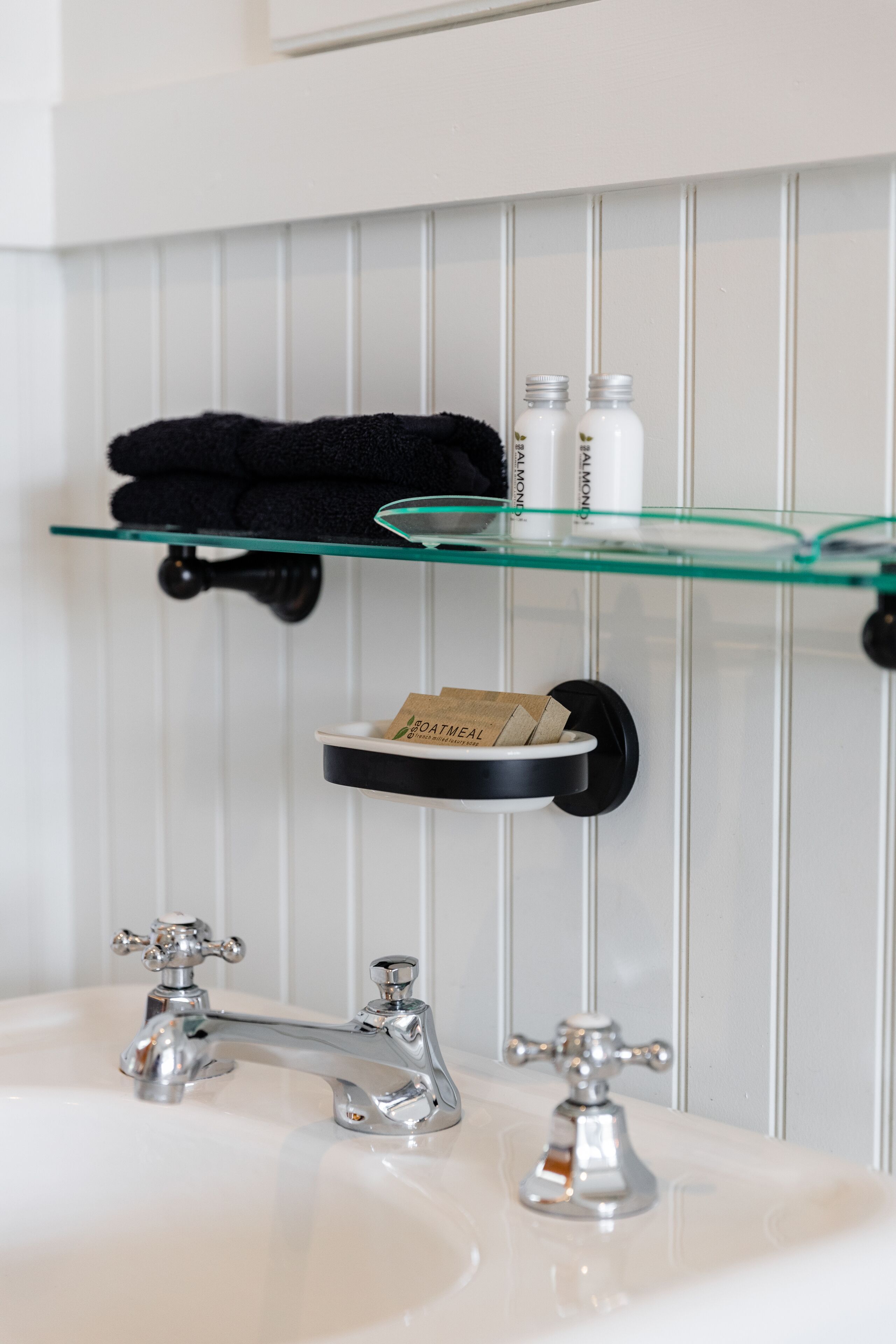 Une salle de bain avec un lavabo blanc, deux robinets, une étagère en verre sur laquelle sont posés une serviette et des articles de toilette, et un porte-savon noir.
