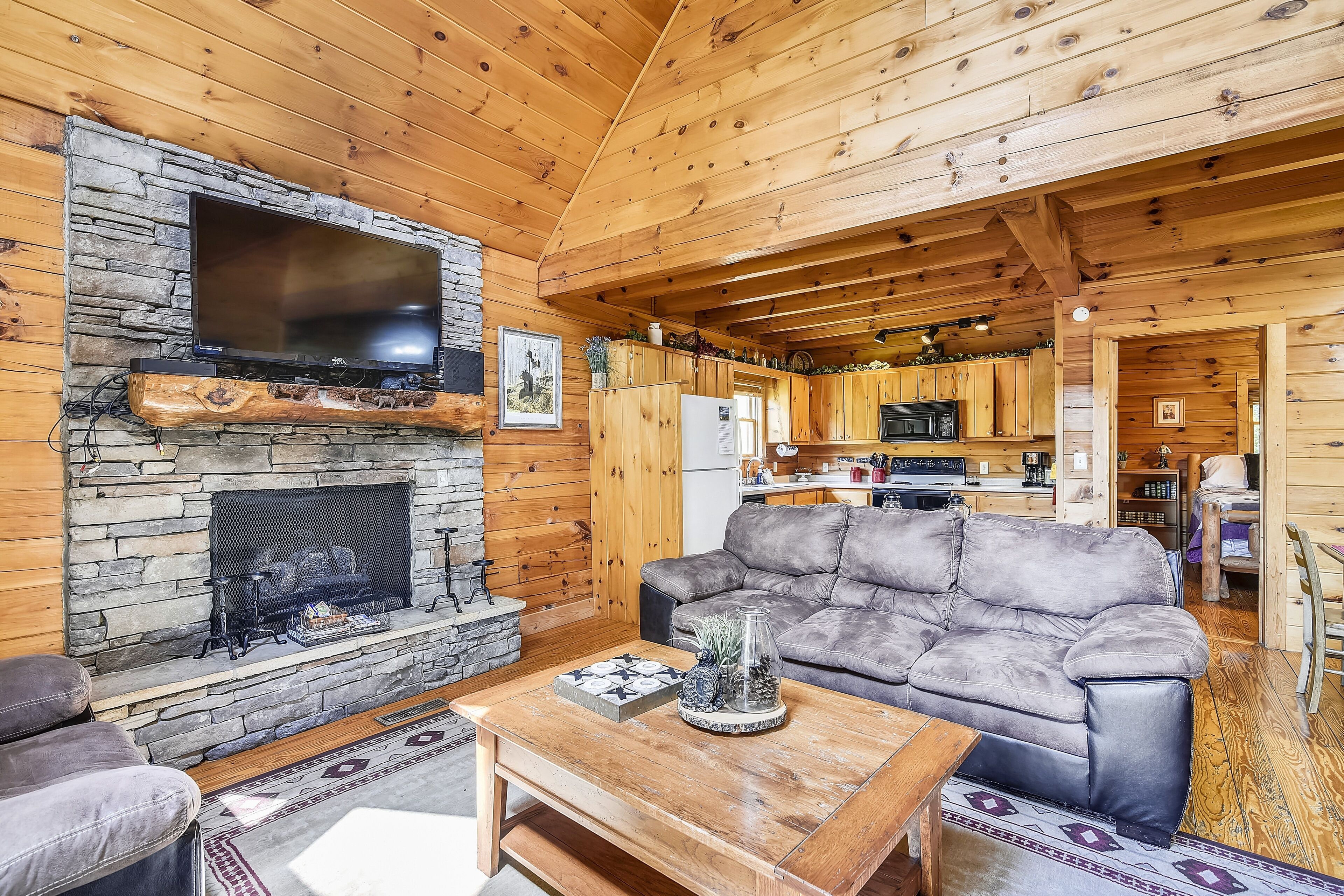 A cozy cabin interior with a stone fireplace, a flat-screen TV, a leather sofa, and a wooden dining table.