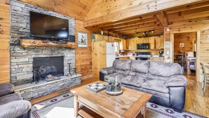 Ein gemütliches Blockhaus-Interieur mit Kamin aus Stein, einem Flachbildfernseher, einer Ledersofa und einem Holzesstisch.