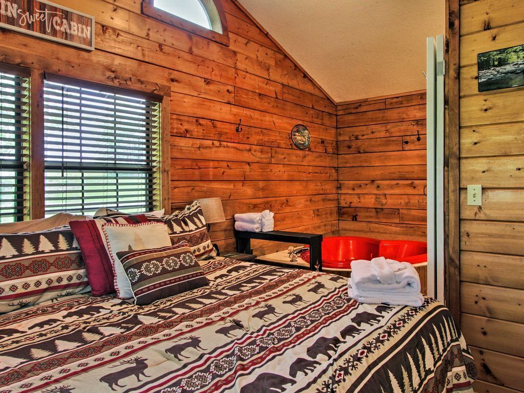 Ein Blockzimmer mit Holzwänden, einem Bett mit gemusterten Betttextilien, einem Fenster mit Jalousien und einem roten Sessel.