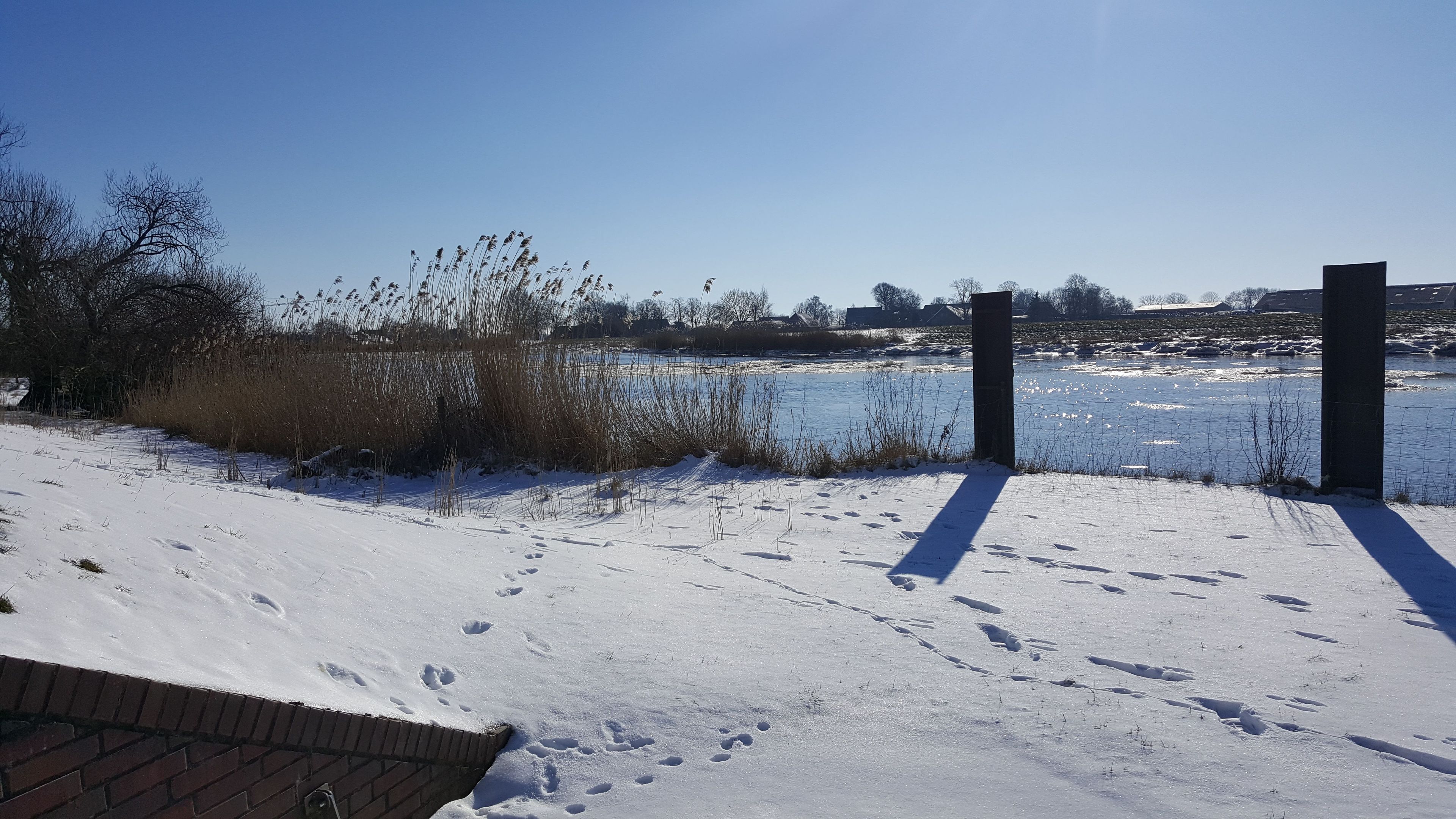 A snowy landscape with a frozen body of water, bare trees, and a brick wall.