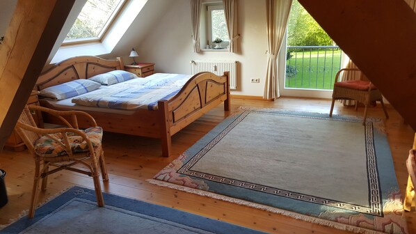 Ein Schlafzimmer mit einem hölzernen Bett, einem Sessel, einem großen Teppich, einem Fenster mit Vorhängen und Blick ins Grüne.