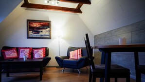 Een knus loft slaapkamer met een schuine kap, een houten tafel, een blauwe fauteuil, een zwarte bank met rode kussens en een lijst met een schilderij aan de muur.