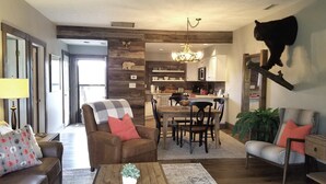 A spacious living room with a brown leather sofa, a wooden coffee table, and a dining area with a wooden table and chairs. A chandelier hangs from the ceiling, and a large black bear sculpture is mounted on the wall.