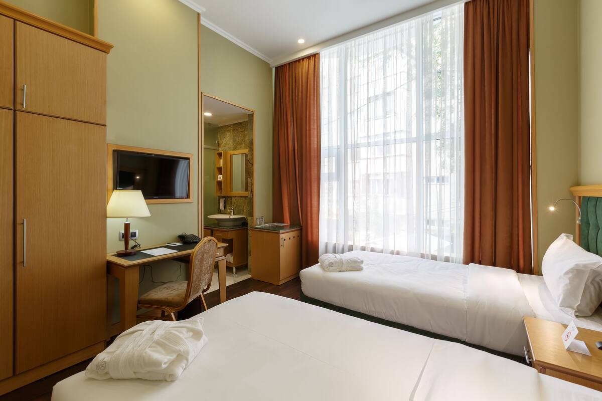 Une chambre d’hôtel avec deux lits, un bureau avec une lampe, un miroir, une salle de bain et de grandes fenêtres avec des rideaux.