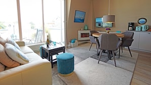 Un salon moderne comprenant un canapé, une table basse et un coin repas avec des chaises et une table. Il y a également un coin cuisine avec une machine à café et un téléviseur.