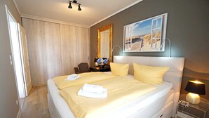 Un lit bien fait, orné d’oreillers jaunes, une table de chevet avec une lampe, un miroir et un tableau représentant une scène de plage.