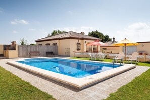 En swimmingpool med flisebelagte omgivelser, en terrasse med udendørs møbler, og en bygning med et flisebelagt tag.