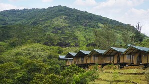 A row of wooden cabins with metal roofs, set against a backdrop of lush green hills. - Wild Glamping (Rangala)