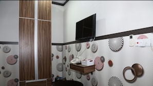 Une salle de bain avec un meuble-lavabo en bois, un miroir, un évier et des toilettes. Un téléviseur est fixé au mur, et une étagère contient des articles de toilette.