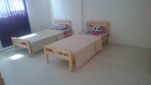 Deux lits simples avec des cadres en bois et une literie beige, disposés dans une pièce aux murs blancs et au sol recouvert d’une moquette.