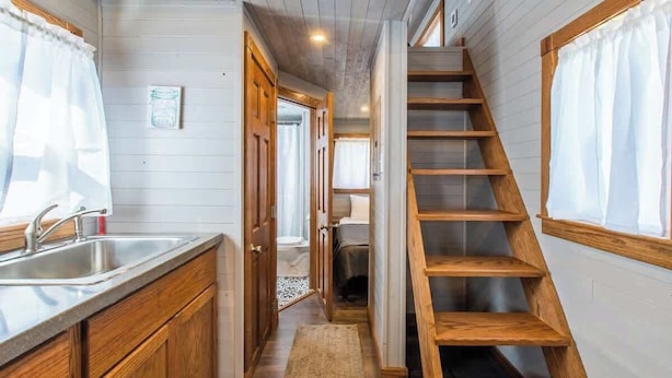 A narrow hallway with a wooden staircase, a kitchen area with a sink and cabinets, and a bed visible in the background.