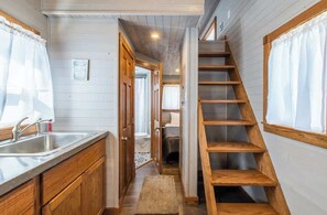 Un estrecho pasillo con una escalera de madera, una zona de cocina con un lavamanos y armarios, y una cama visible al fondo.