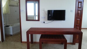 A room with a wooden table, a television mounted on the wall, a mirror, and a shower area. - Hotel Com Apartment (Entebbe)