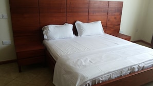 A bedroom with a wooden headboard, a large bed with white bedding, and a nightstand. - Hotel Com Apartment (Entebbe)