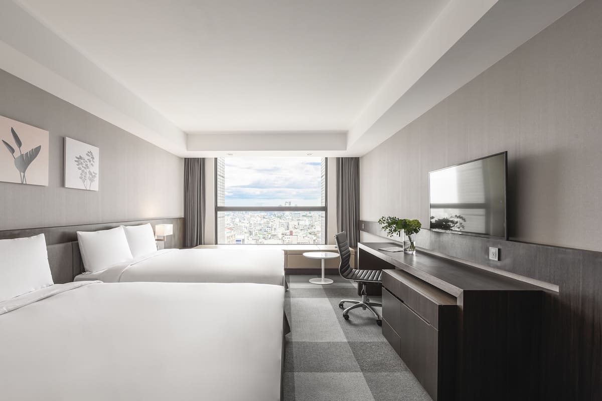 A modern hotel room with two beds, a desk, a chair, a TV, and a view of the city.