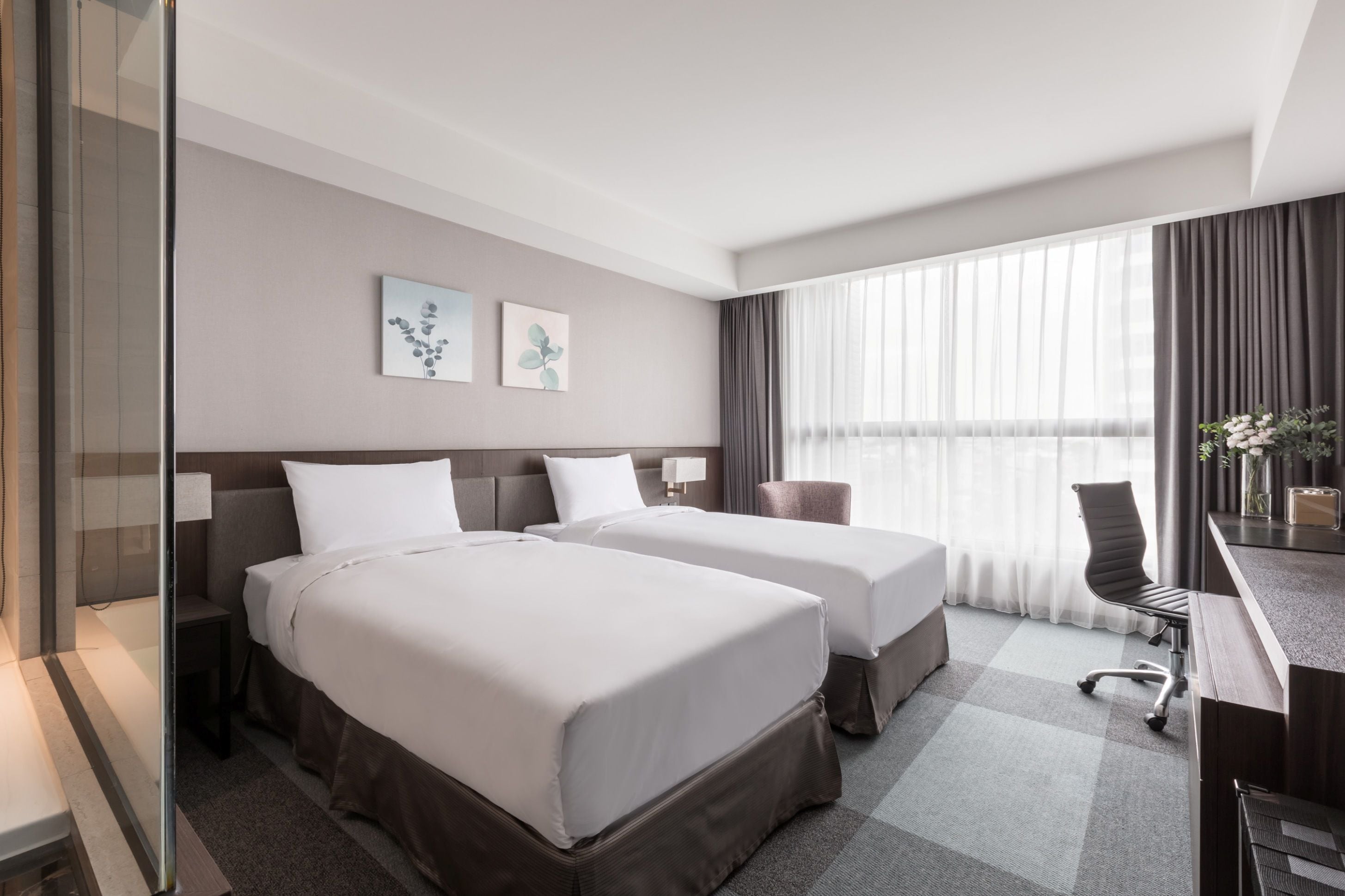 A hotel room with two beds, a desk, a chair, and a large window with curtains.