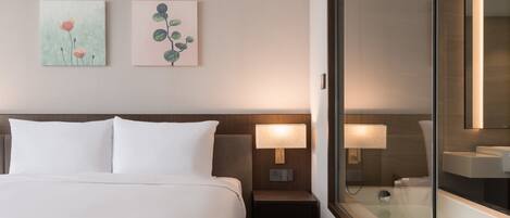 Habitación de hotel con cama, mesitas de noche, lámpara, cabina de ducha de vidrio y dos obras de arte colgadas en la pared.