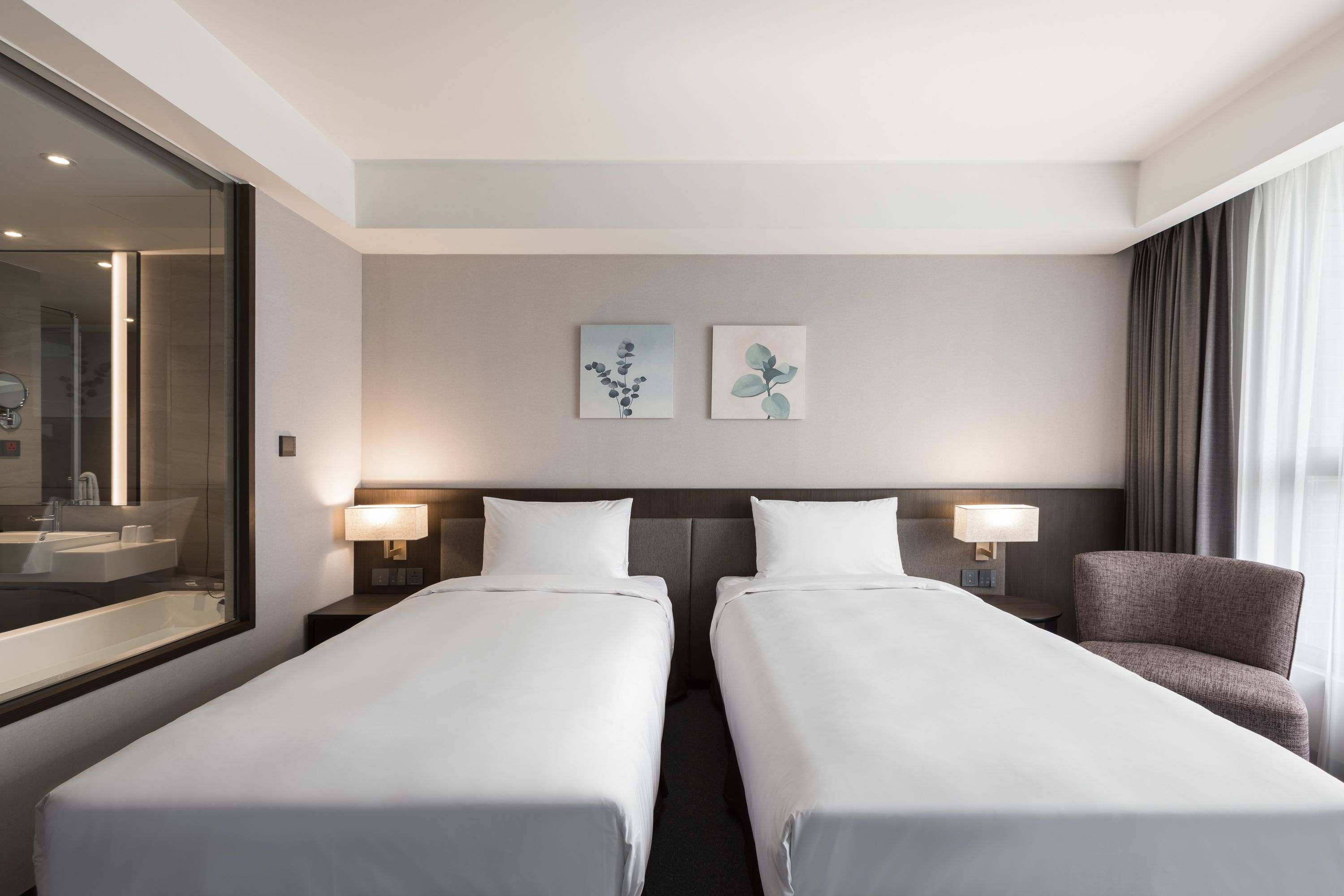 A hotel room with two single beds, a large mirror, a bathroom with a sink and mirror, and two wall-mounted lamps.