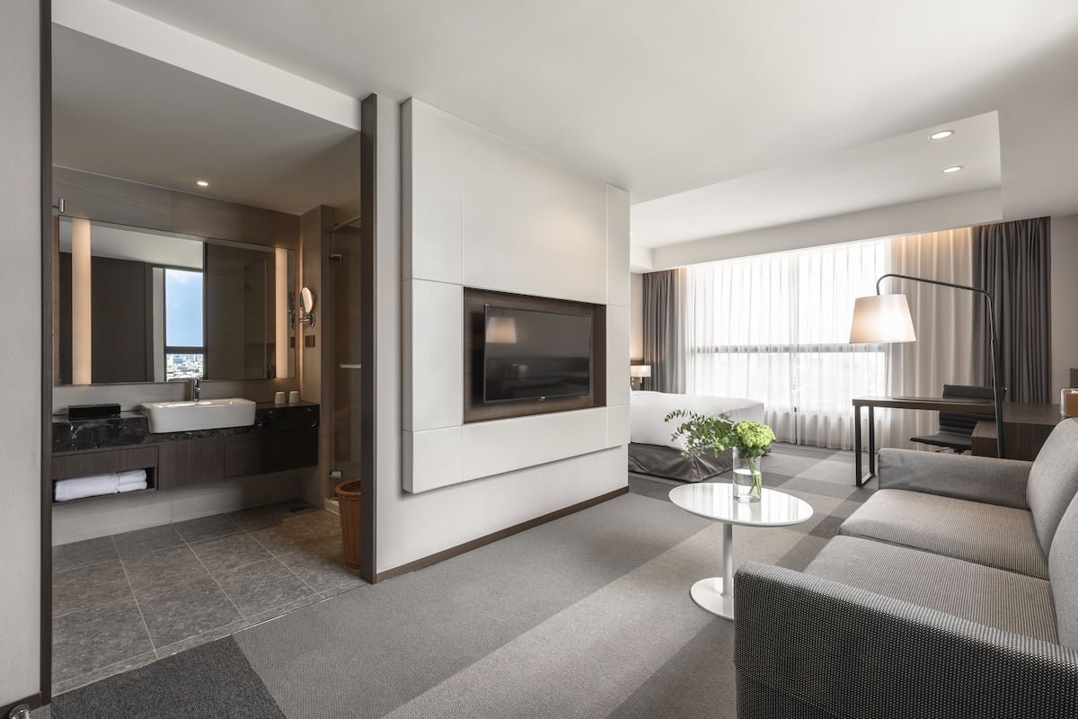 A modern hotel room with a flat-screen TV mounted on the wall, a sofa, a coffee table, and a large window with curtains.