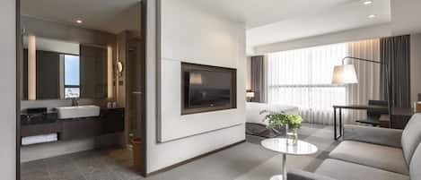 A modern hotel room with a flat-screen TV mounted on the wall, a sofa, a coffee table, and a large window with curtains.