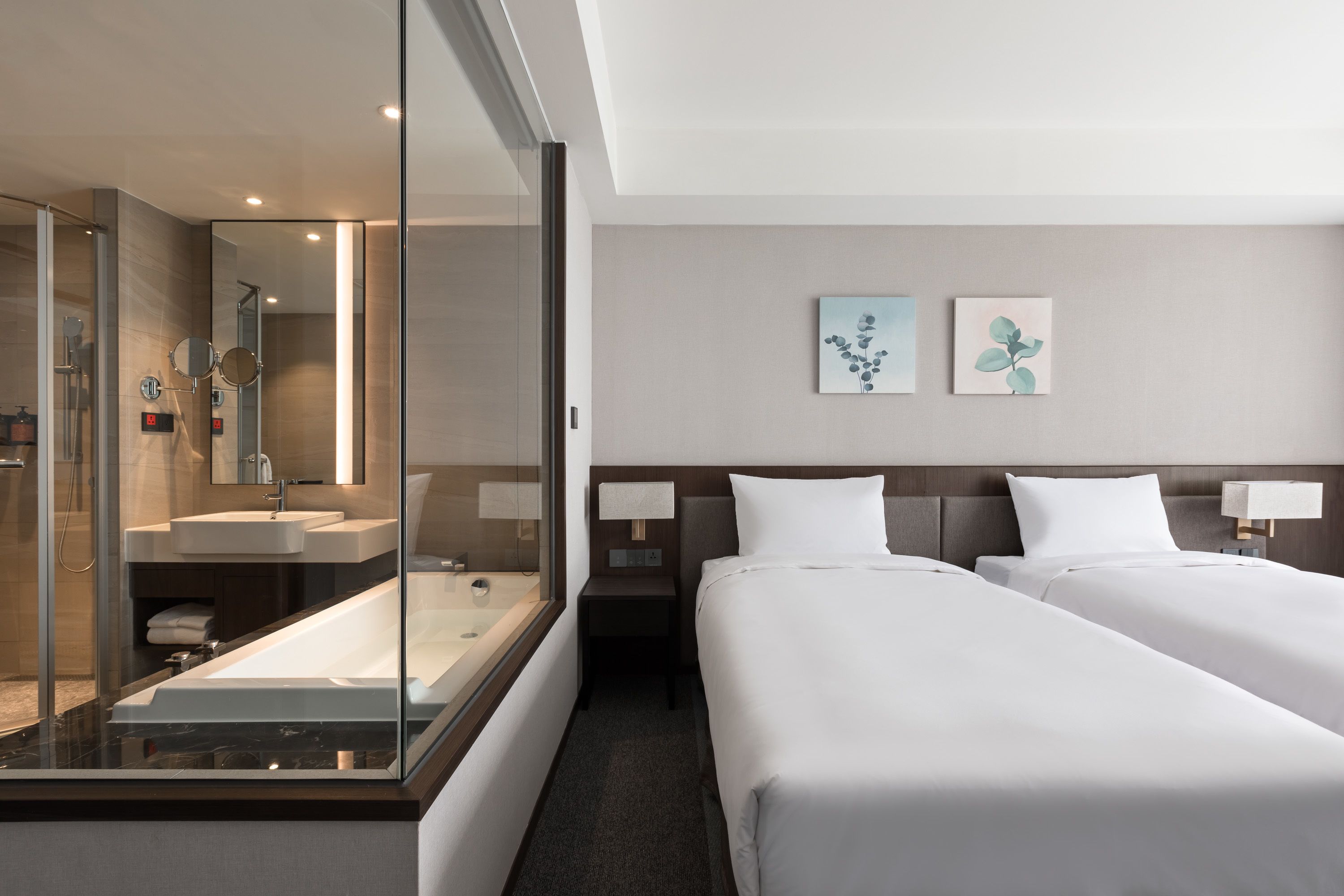 Quarto de hotel com chuveiro em vidro, uma cama de casal, mesa de cabeceira com abajur e duas pinturas penduradas na parede.