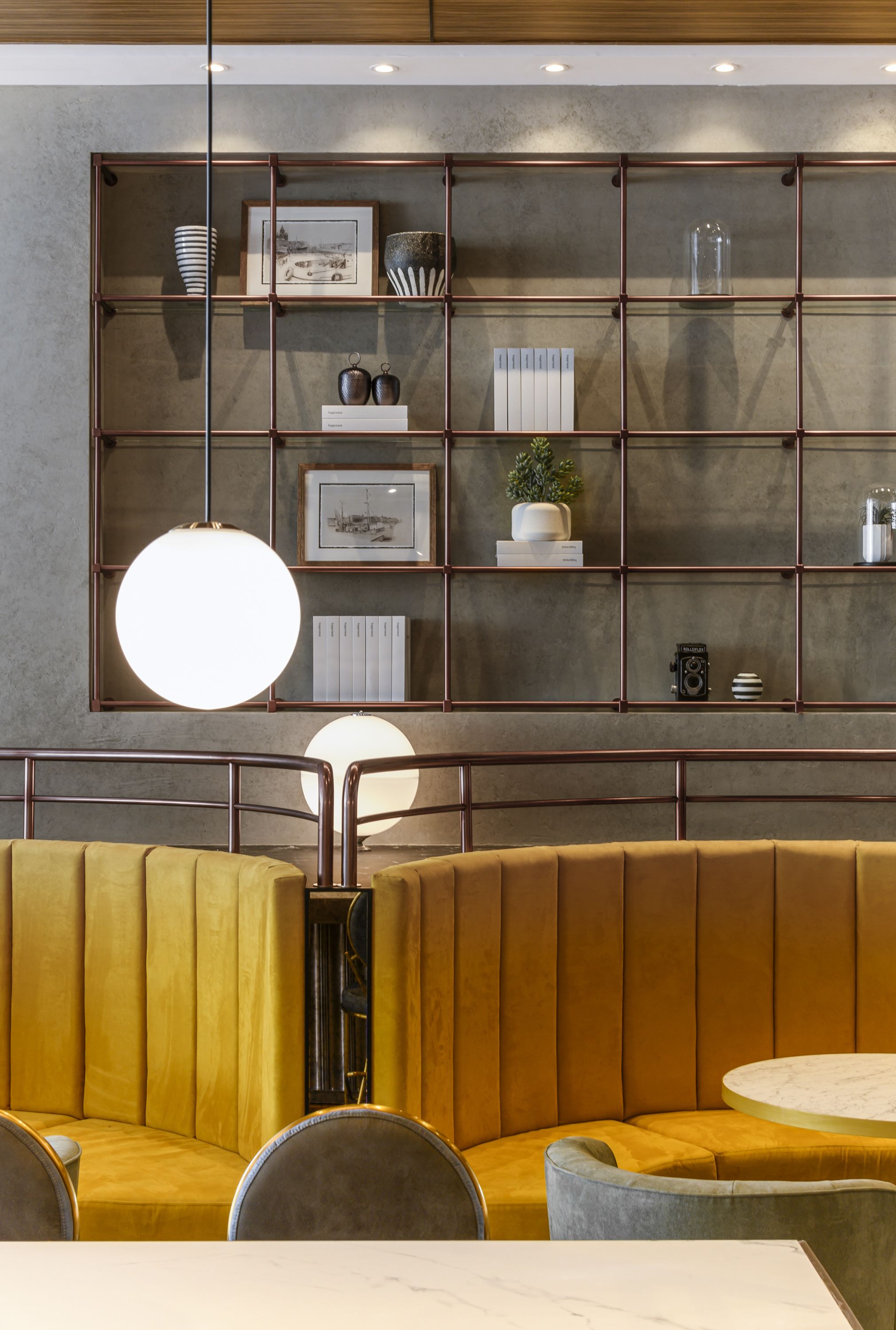 A modern interior with a seating area featuring a yellow booth and a round table. A metal-framed shelving unit with various decorative items is mounted on the wall.