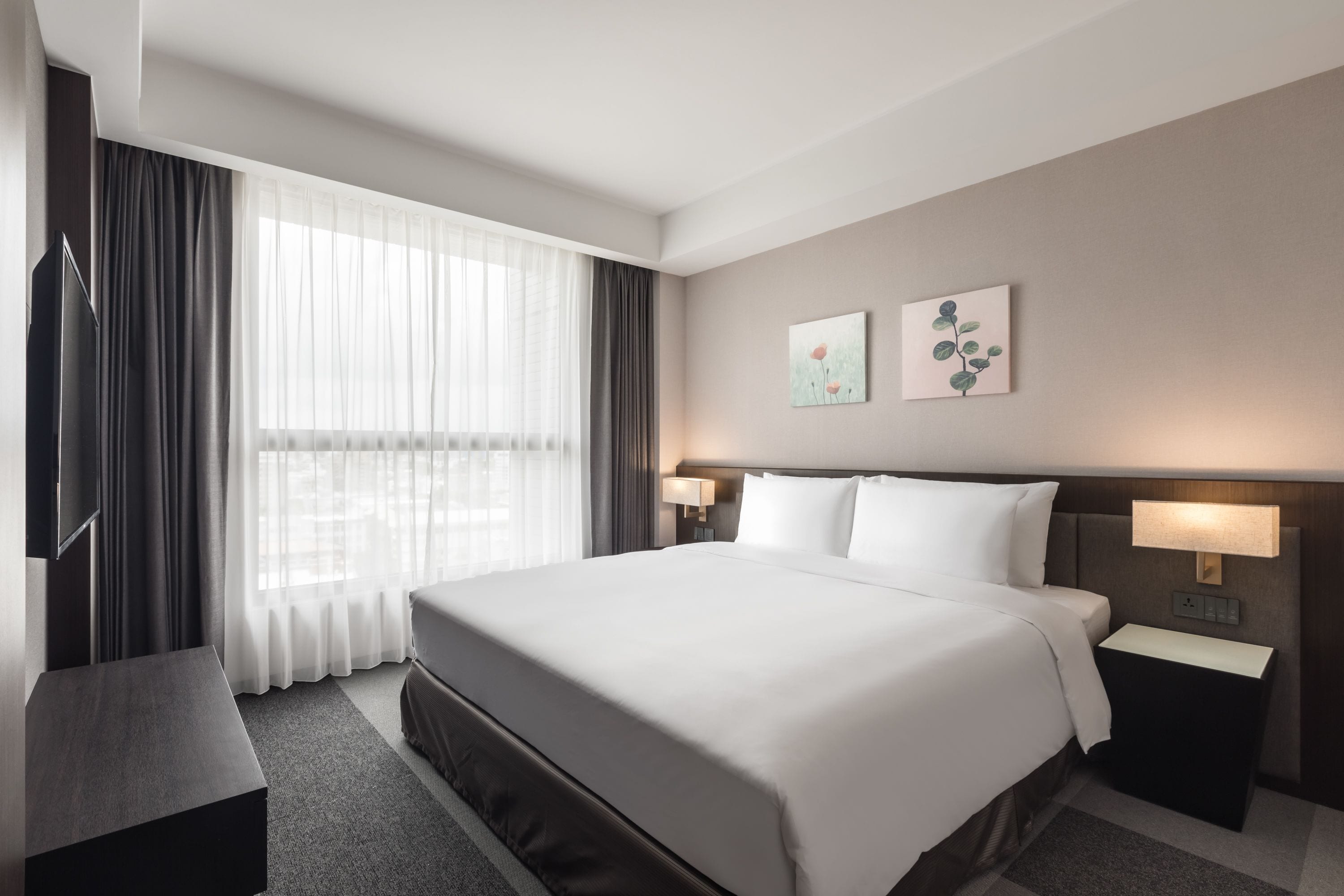 A modern hotel room with a large bed, two bedside lamps, a flat-screen TV, and a large window with sheer curtains.