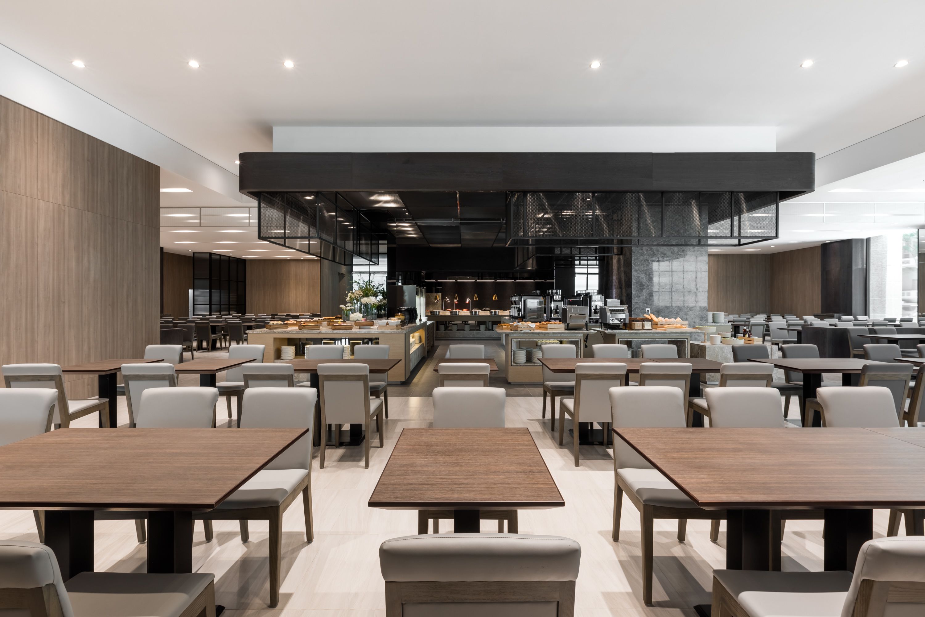 A spacious dining area with multiple tables and chairs, a central kitchen area, and a modern architectural design with exposed ceilings and natural light.