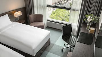 A hotel room with a bed, a chair, a desk, a lamp, a vase with flowers, and a window with a city view.