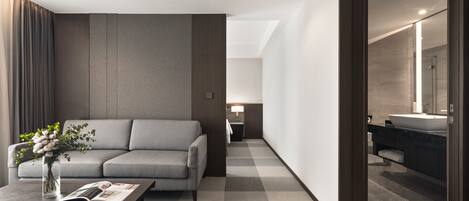 Una habitación de hotel moderna con un sofá gris, una mesa de centro con una revista, un jarrón de flores y vistas al baño con un lavabo y espejo.