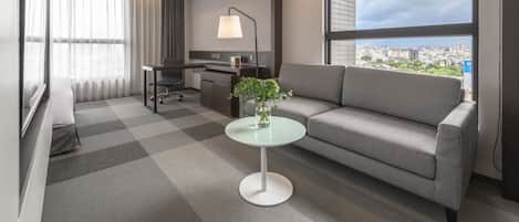 Een moderne hotelkamer met een bank, een kleine tafel met een bloemenvaas, een bureau met een lamp en een groot raam met uitzicht op de stad.