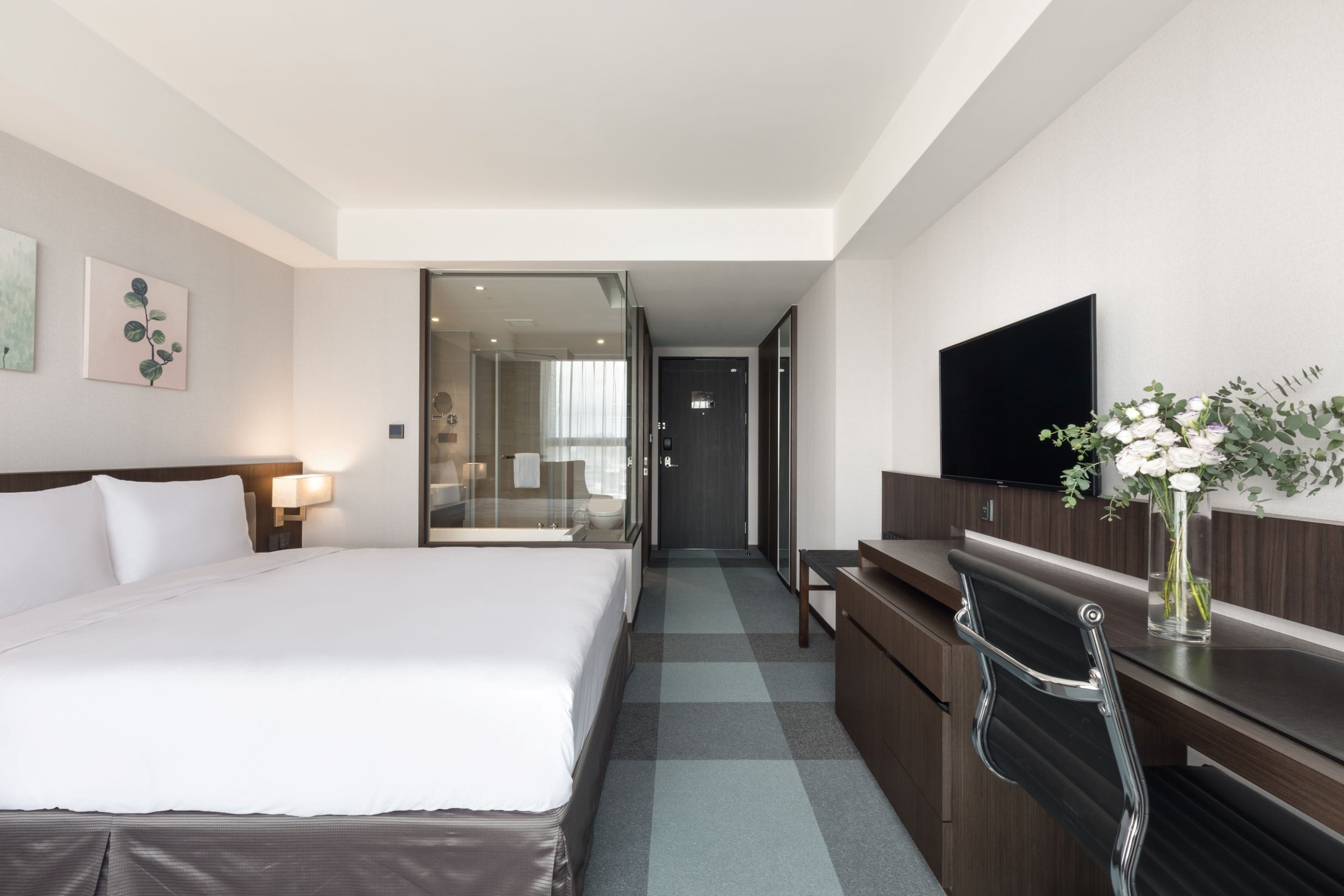A hotel room with a large bed, a desk, a chair, a TV, and a vase with flowers.