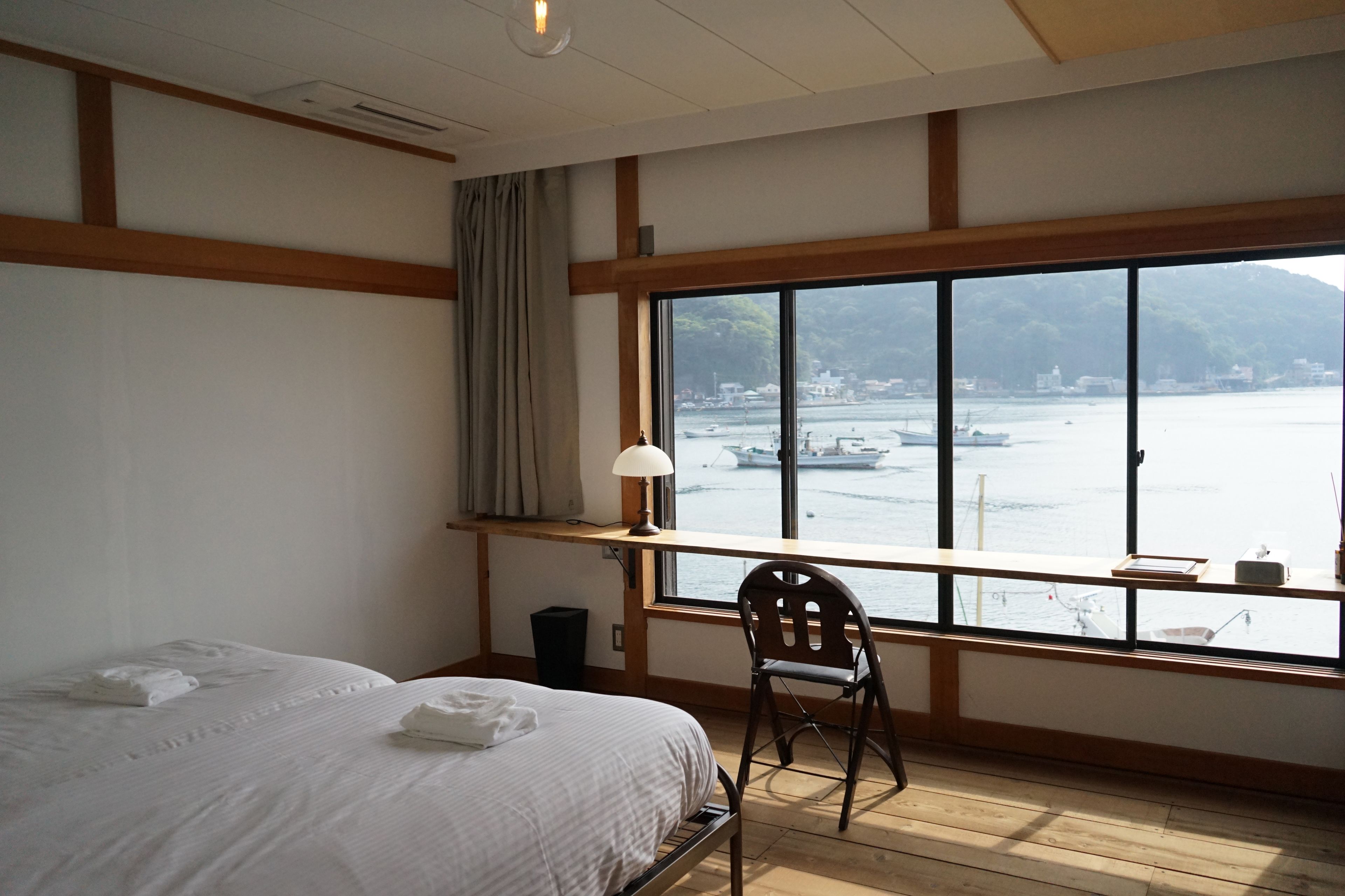 A hotel room with a large window, a bed, a chair, and a desk with a lamp.