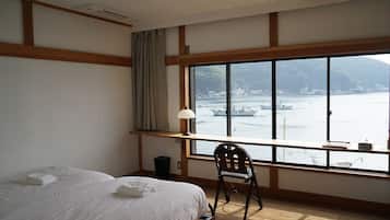 A hotel room with a large window, a bed, a chair, and a desk with a lamp.