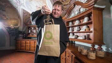A person holding a paper bag with a logo, standing in a room with ornate ceiling and shelves of ceramic items.