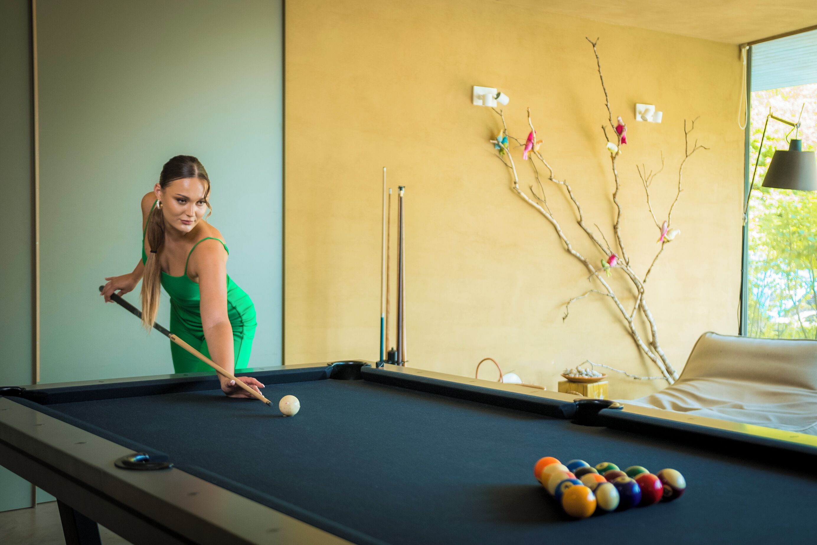 A pool table with balls and a cue stick, a decorative branch with ornaments, and a bed with a lamp in a room with a window.