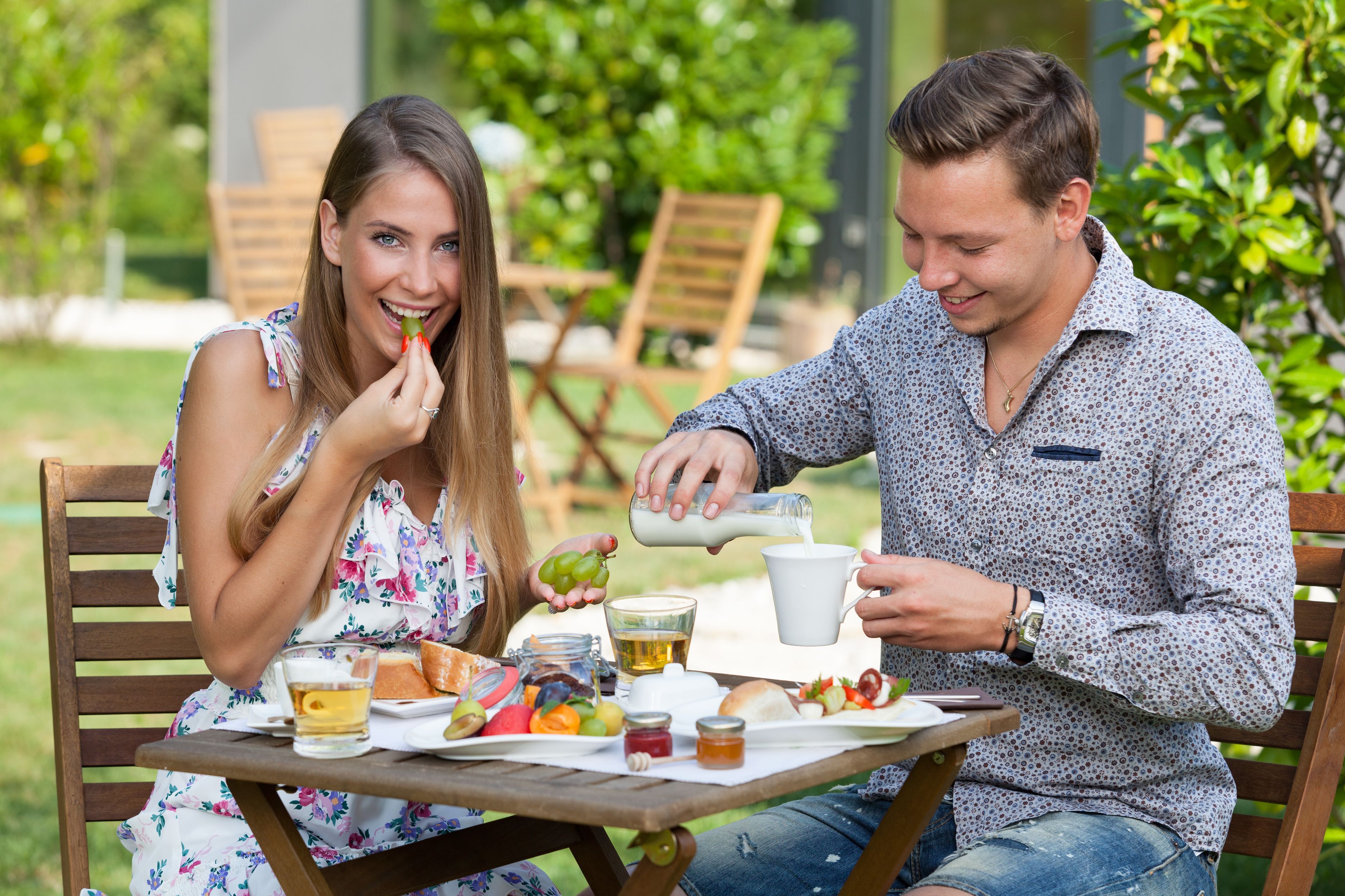 A couple dining outdoors at a wooden table with a meal, including bread, fruits, and a beverage.